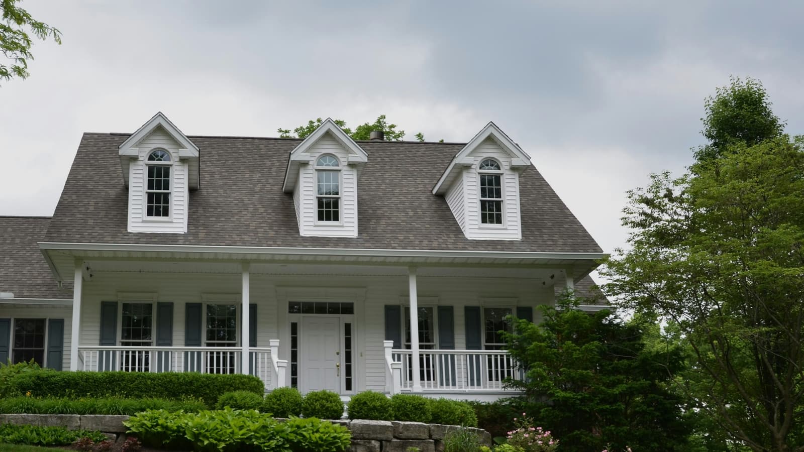 White Cape Cod style house with dormers and a front porch behind a landscaped green lawn